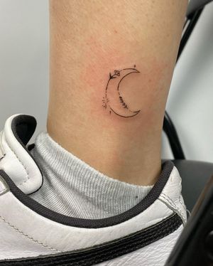 Achieve delicate beauty with this ankle tattoo featuring a moon and flower design, created by expert artist Tianna.