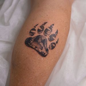 Capture the spirit of the wilderness with this blackwork lower leg tattoo featuring a majestic bear, towering mountains, a towering tree, and a bold paw print. By artist Yasmin Clara.