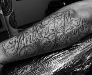 This stunning blackwork forearm tattoo by Lokey features intricate lettering and illustrative design, showcasing a name and quote.