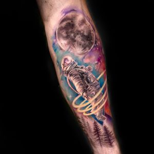 Capture the magic of space with this illustrative watercolor tattoo featuring a moon, astronaut, and tree on your forearm.