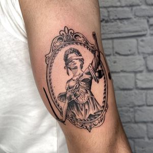 Experience the balance of justice and strength in this stunning upper arm tattoo by Marcos. Featuring a powerful woman holding a sword framed by intricate filigree.