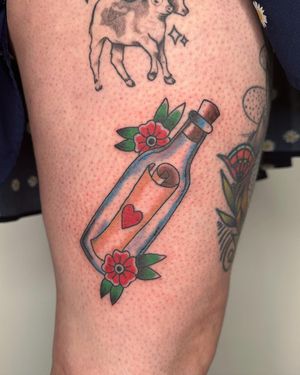 A beautiful illustrative tattoo on the upper leg featuring a flower, heart, bottle, and letter motif, done by talented artist Sasha.