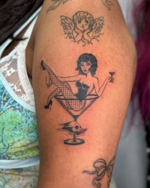 This blackwork fine line tattoo on the upper arm features a skull, woman, glass, skirt, and drink in a stylish illustrative design.