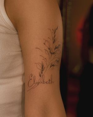Beautiful fine line tattoo of a flower with small lettering of a name on upper arm by Fabian Lopez Barreda.