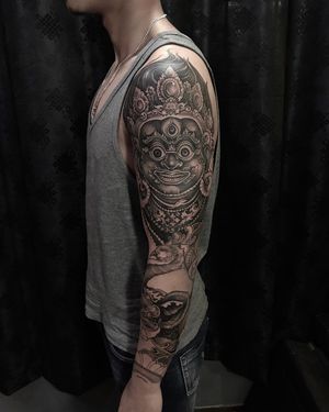 Elegant black and gray sleeve tattoo featuring a stunning Hindu mask design, expertly crafted by the talented artist Avi.