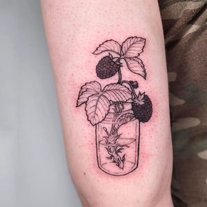 This upper arm tattoo features a stunning design of flowers, fruits, bottles, and leaves, beautifully rendered in blackwork style by the talented artist Erin.