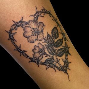 A stunning black & gray forearm tattoo featuring a delicate flower entwined with barbed wire by Letitia Mortimer.