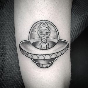 A black and gray tattoo on the upper arm featuring a ship, alien, and spaceship by Matthew Ono. Perfect for sci-fi enthusiasts!
