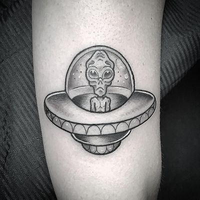 A black and gray tattoo on the upper arm featuring a ship, alien, and spaceship by Matthew Ono. Perfect for sci-fi enthusiasts!