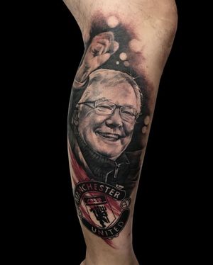 An illustrative black and gray tattoo on the lower leg, featuring a man in glasses with a soccer motif, by Avi.