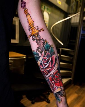 Unique forearm tattoo with small lettering by Frankie Brown blending a flower and sword motif.