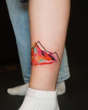 Discover a stunning lower leg tattoo featuring a mountain motif, beautifully executed in geometric and watercolor styles by the talented artist Aygul.