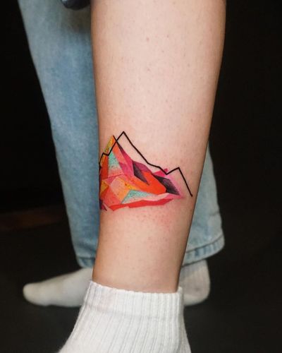 Discover a stunning lower leg tattoo featuring a mountain motif, beautifully executed in geometric and watercolor styles by the talented artist Aygul.