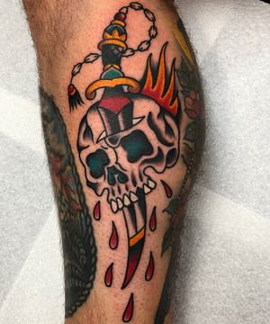 Get a bold and classic tattoo featuring a skull and dagger design on your lower leg, executed by the talented artist Carlos Zucato.