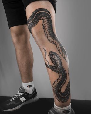 Experience the stunning artistry of FKM TATTOO with this illustrative snake tattoo, elegantly placed on the knee in bold blackwork style.
