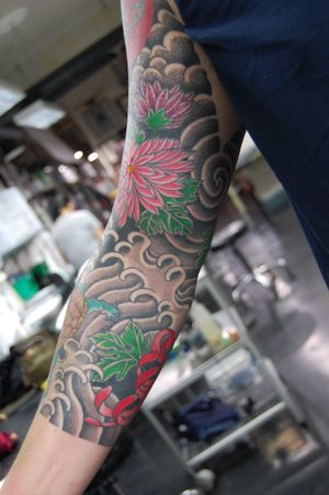 Experience the beauty of Japanese art with this stunning arm tattoo featuring intricate floral motifs and swirling waves by the talented artist Bananajims.