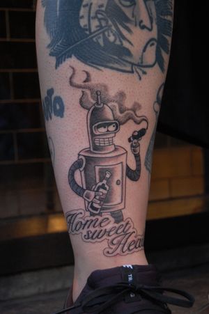 Check out this cool black and gray lettering tattoo on the lower leg, featuring a robot and a bottle inspired by Futurama, done by Bananajims.