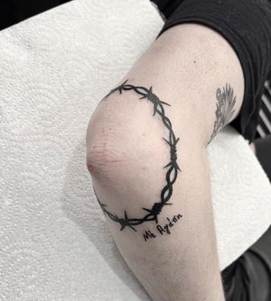 Bold blackwork design by Federico Colantoni combining barbed wire motif and meaningful quote on the knee.