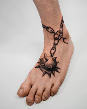 Detailed black and gray tattoo by Delphin Musquet featuring a chain and spike motif on the foot.