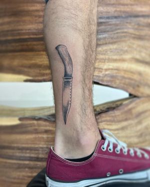 Check out this edgy blackwork knife tattoo by Hana Kaki, located on the lower leg for a unique and bold look.