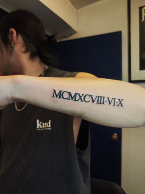 A stylish lettering tattoo featuring Roman numerals and numbers on the forearm, created by the talented artist Nikki Bostin.