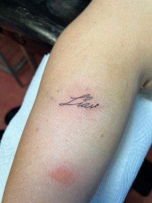 Get a beautiful and intricate fine line name tattoo on your upper arm by Kayleigh Cole. Perfect for a subtle and personalized touch.
