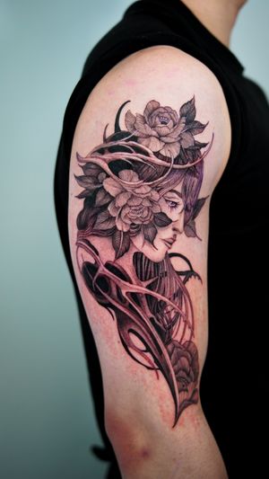 Explore the intricate details of this stunning black and gray tattoo on the upper arm, featuring a beautiful woman, mesmerizing eyes, and delicate flower elements. A fusion of neo-traditional and realism styles.