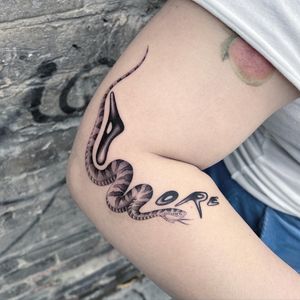 A captivating black and gray tattoo featuring a snake entwined with small lettering on the arm, crafted by artist Fresh Flower.