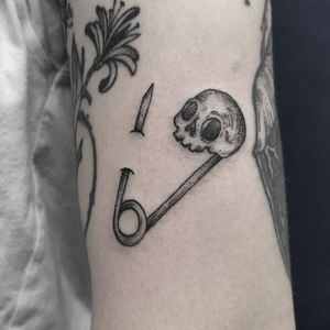 Black and gray tattoo featuring a skull and pin design by Elisa Thirteen. Bold and unique arm placement.