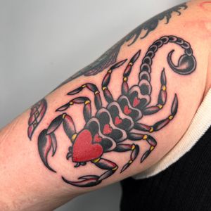 Get a fierce and loving tattoo with a traditional style scorpion and heart motif on your upper arm by the talented artist Claudia Trash.