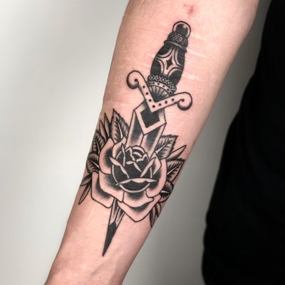 Adorn your forearm with a stunning traditional tattoo featuring a vibrant rose and sharp dagger, expertly executed by Claudia Trash.