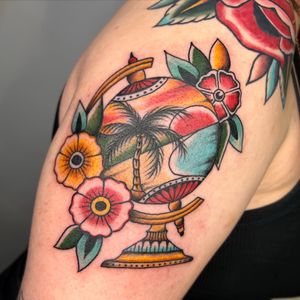 A stunning traditional tattoo featuring a beautiful beach scene intertwined with delicate flowers, expertly done by the talented artist Claudia Trash on the upper arm.