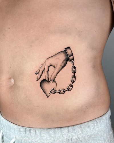 Unique black and gray tattoo by Joshua Williams featuring a heart held in a hand connected to a chain. Perfect for the ribs.