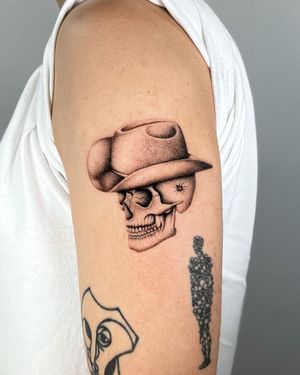 Black and gray tattoo of a skull wearing a hat on the upper arm, done by tattoo artist Joshua Williams.