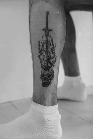 Get a bold blackwork tattoo of a skull and sword on your lower leg by Jamie B for a powerful and edgy look.