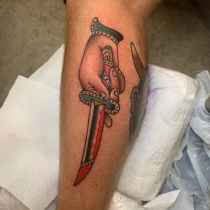 Get inked with a bold traditional tattoo by Andrea Furci featuring a dagger and hand design on your lower leg.