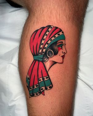 Check out this classic traditional tattoo featuring a woman wearing a bandana on the lower leg, inked by the talented artist Ryan Goodrum.