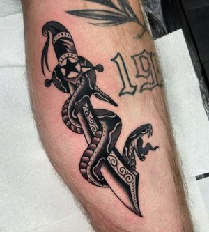 Get inked by Ryan Goodrum with this classic design featuring a menacing snake and dagger on your lower leg. Stand out with this bold traditional piece!