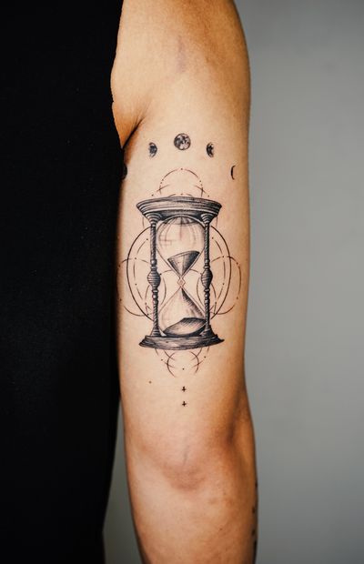 Explore the timeless beauty of a black & gray fine line tattoo featuring a moon and hourglass design by Gabriele Edu on your upper arm.