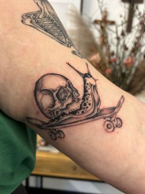 A unique black and gray, fine line tattoo featuring a skull, skateboard, and snail motif by artist Jack Howard.