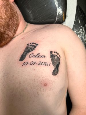 Intricate black and gray baby feet print with small lettering of name and date, beautifully crafted on the chest by tattoo artist Kiky Flore.