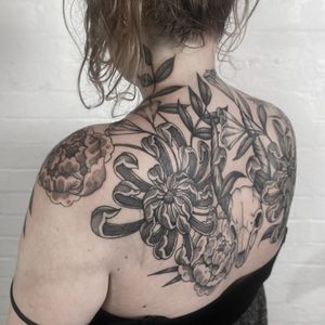 A black and gray floral tattoo on upper back featuring a chrysanthemum, skull, horns, and leaves. Designed by Jenny Dubet.