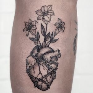 A traditional black and gray tattoo on the lower leg, featuring a detailed anatomical heart intertwined with delicate floral elements. Created by Jenny Dubet.