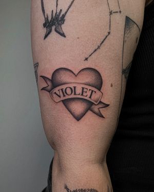 Get a timeless traditional tattoo of a heart with a custom name by Ophelya Jeandat on your arm.