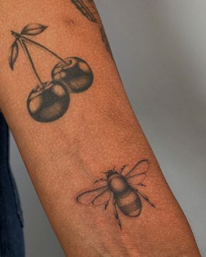 Stunning black and gray tattoo featuring a detailed bee and cherry motif, created by the talented artist Ophelya Jeandat.