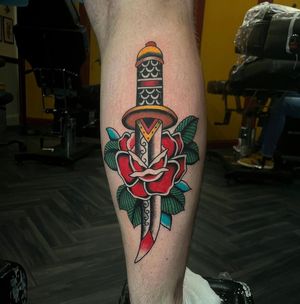 Get inked with a bold traditional style tattoo featuring a stunning rose and sharp dagger design by Barney Coles.