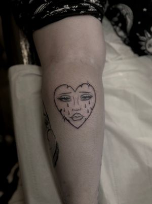 Illustrative traditional tattoo featuring a sad face within a heart motif, beautifully crafted by Maddie.