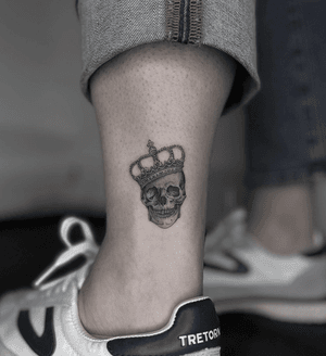 Get inked with a stunning black and gray skull and crown design on your lower leg by tattoo artist Kayla. Make a bold statement with this regal and edgy piece!