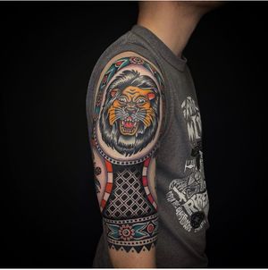 Get inked with a fierce lion surrounded by intricate patterns and a ribbon in traditional tattoo style by artist Sam Young. Roar with pride! 