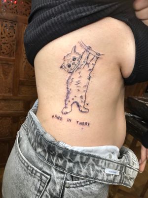 Small lettering and illustrative tattoo of a cat hanging on a tree by artist Jonathan Glick. Full of ignorant charm.